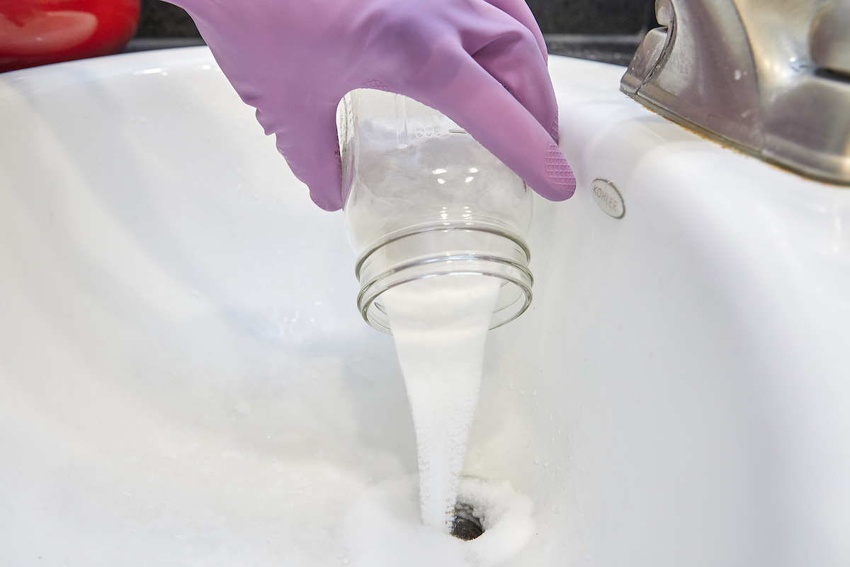 Woman wearing purple rubber gloves pours homemade drain cleaner from a mason jar into a bathroom sink.