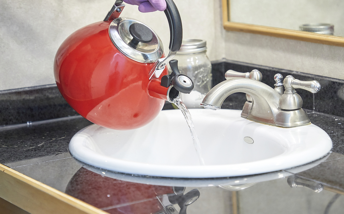 Woman uses red tea kettle to pour hot water down a bathroom sink.