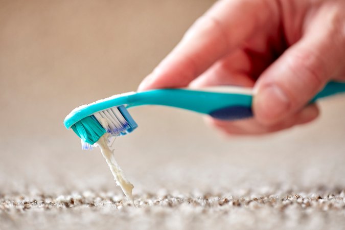 Close up of person holding an aqua toothbrush pulling chewing gum out of a beige carpet.