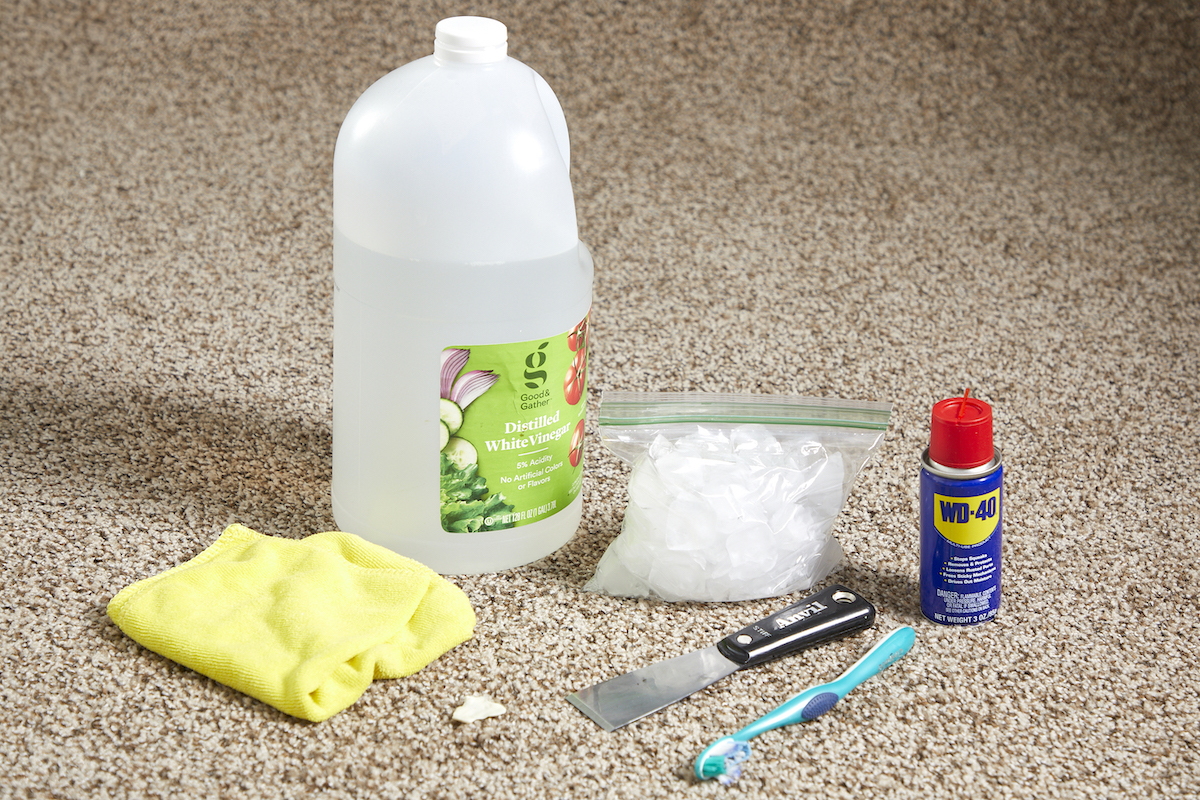 Various cleaning materials laid on a beige carpet, including WD-40, bottle of vinegar, rag, ice, and toothbrush.