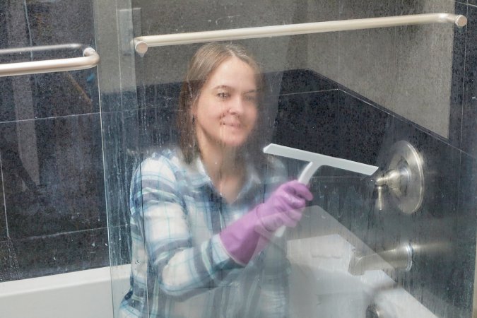 Woman crouches inside her bathtub behind dirty shower door, cleaning it with a squeegee.