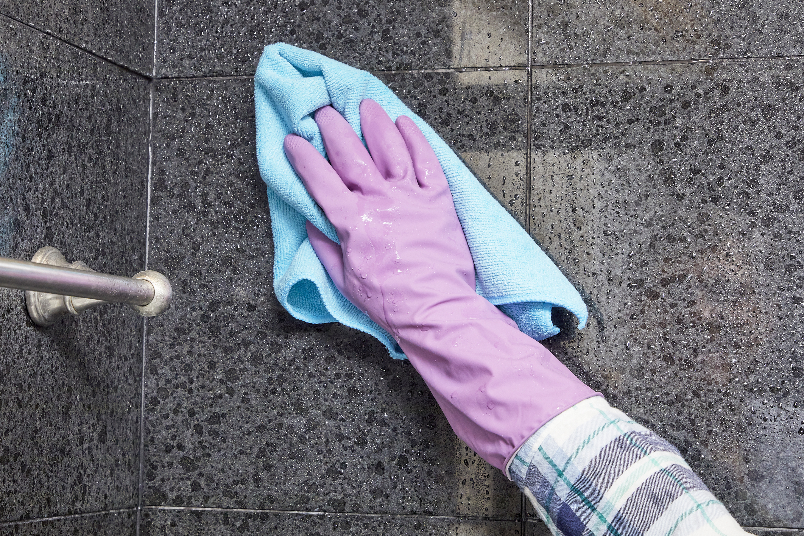 Woman wearing purple rubber gloves dries the tile walls of her bathtub with a microfiber cloth.