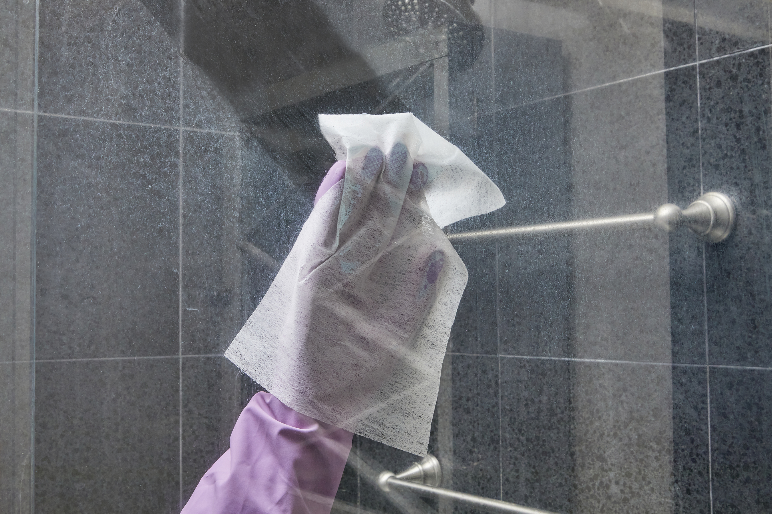 Woman wearing purple rubber gloves wipes shower door with a dryer sheet.