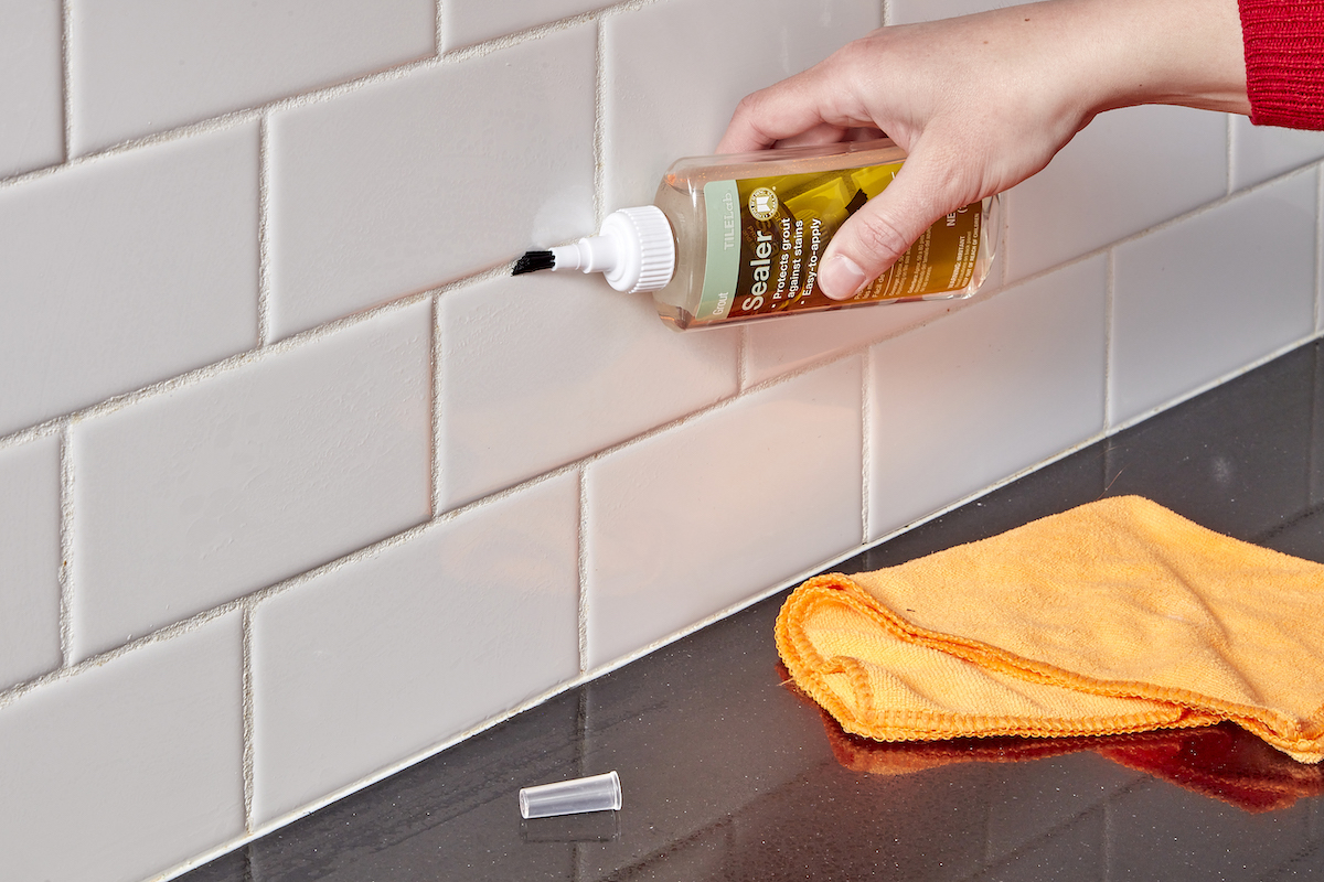 Woman uses small bottle of grout sealant to paint grout between tiles.