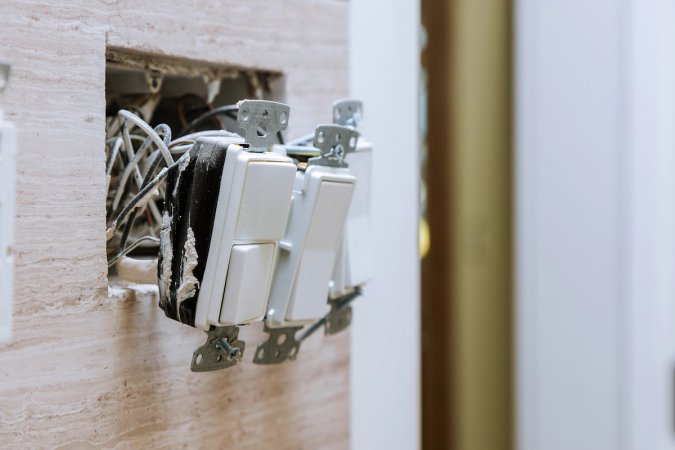 Two adjacent light switches with two toggles each are hanging from the wall via their wires.