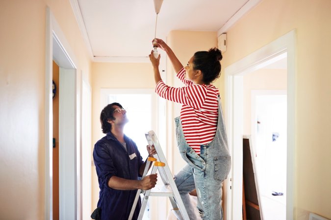 A pregnant woman wearing overalls stands on a ladder held steady by a younger man to change an interior lightbulb.