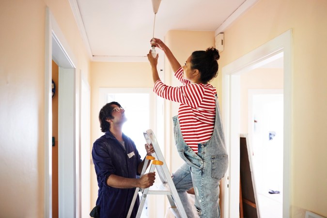 A pregnant woman wearing overalls stands on a ladder held steady by a younger man to change an interior lightbulb.