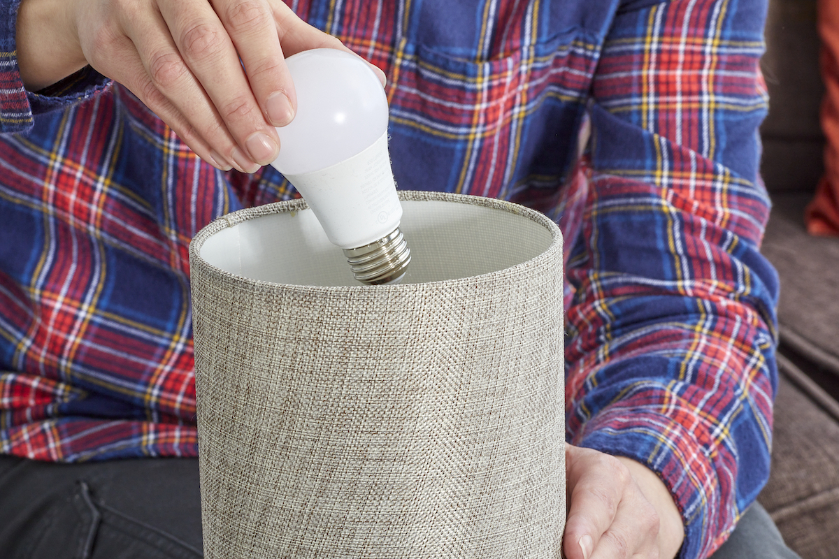 Woman wearing a plaid flannel shirt removes a light bulb from a small lamp with a beige shade.