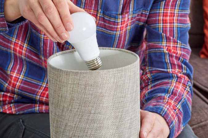 Woman wearing a plaid flannel shirt removes a light bulb from a small lamp with a beige shade.