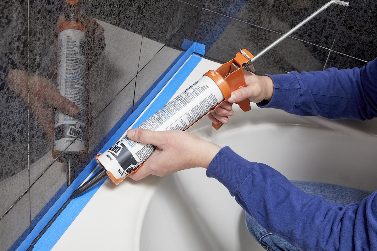 Woman uses a caulking gun to caulk the gap between the bathtub and the tile wall.