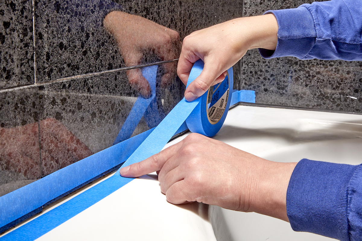 Woman uses painter's tape to protect tub and wall from caulk application.