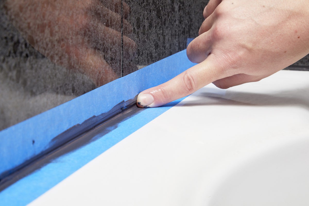 Woman uses her finger to smooth the bead of caulk; painter's tape keeps it off the tile.