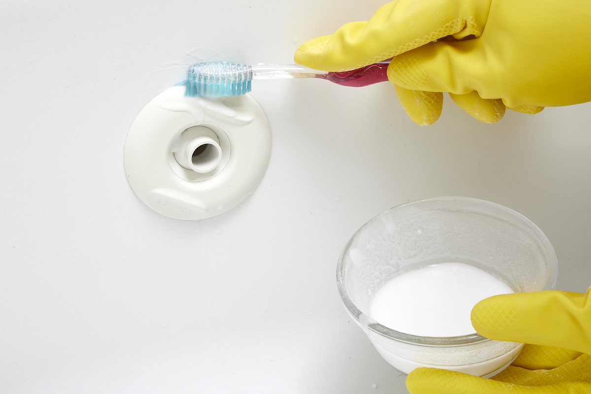 Woman wearing yellow rubber gloves and holding small bowl with baking soda mixture cleans tub jets with a toothbrush.