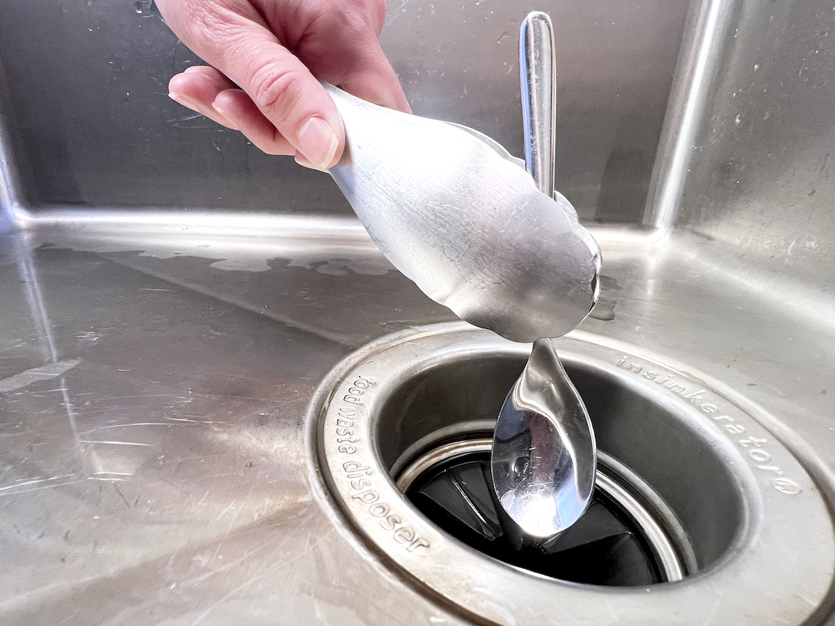 Woman uses tongs to pull spoon out of a garbage disposal.