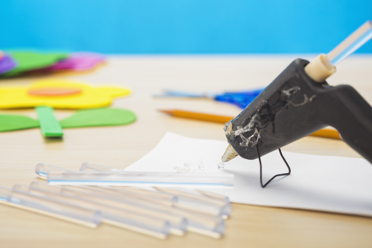 A mini hot glue gun with a blob of clear glue oozing out of the tip onto a scrap piece of white paper, with scissors and a pencil, along with a yellow flower created from foam sheets on a light colored wood grain table and a blue wall in the background.