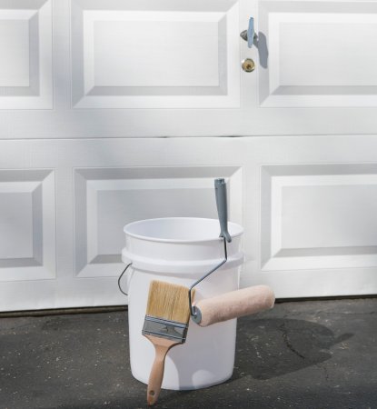 A paint brush, roller and bucket in front of a white garage door.