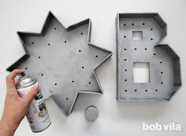 A person spray paints the marquee letter with a top coat of hammered silver.