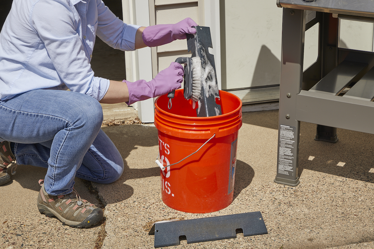 Woman washes grill parts in soapy water in a 5-gallon bucket.