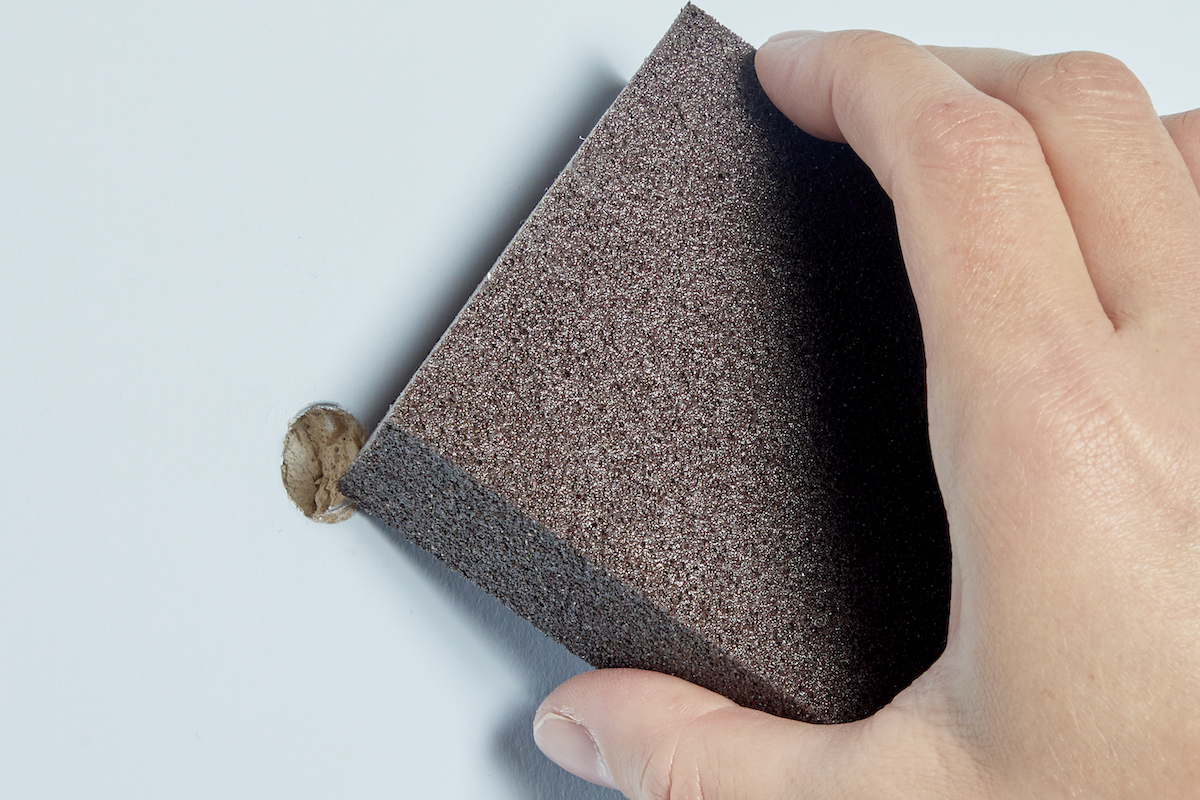 Woman uses a sanding block to clean a hole in drywall. 