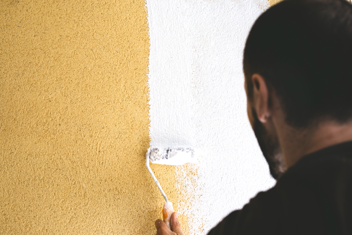 Back view of man painting a yellow stucco wall white.