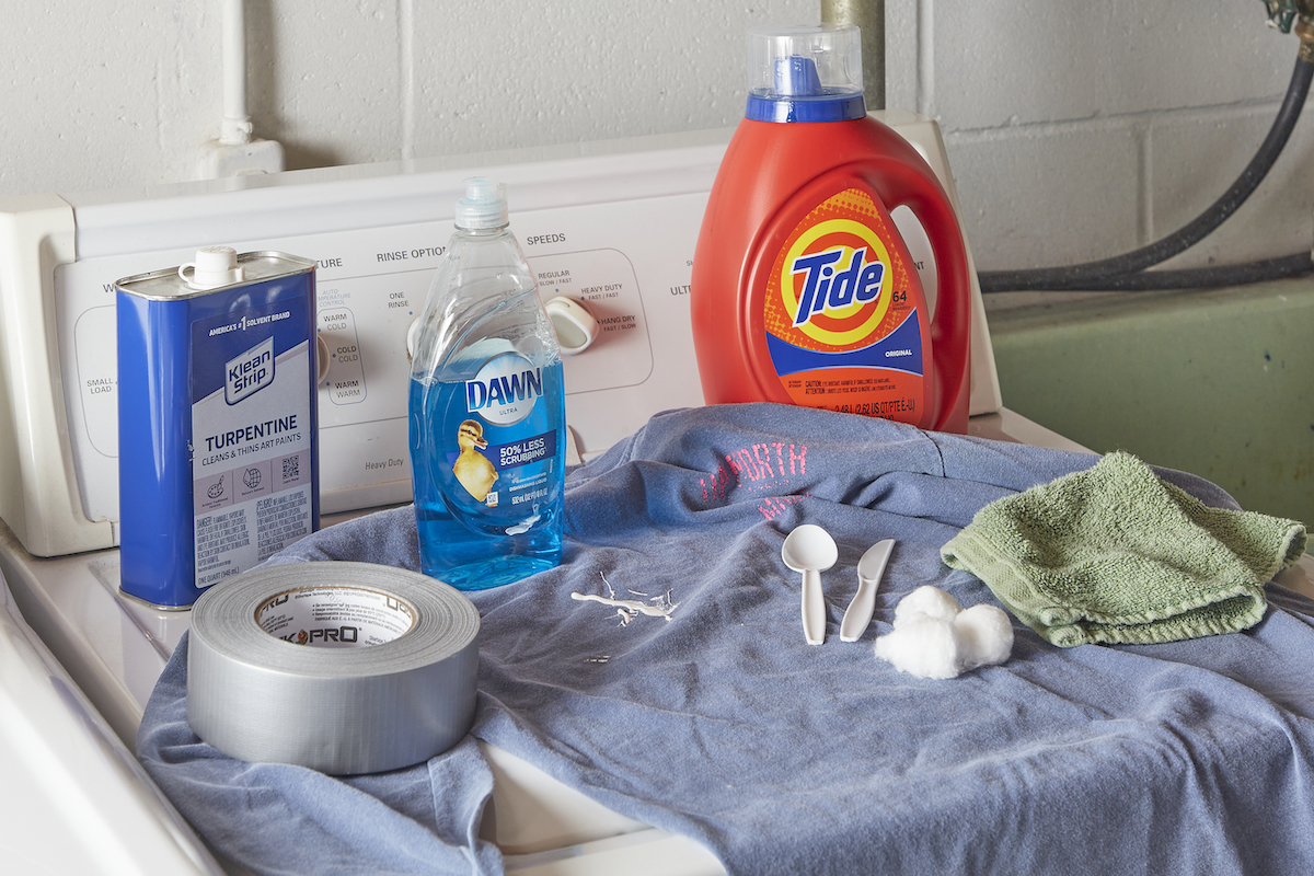 On top of a top-load washing machine, a paint-spattered shirt and the materials needed to remove paint from the shirt.