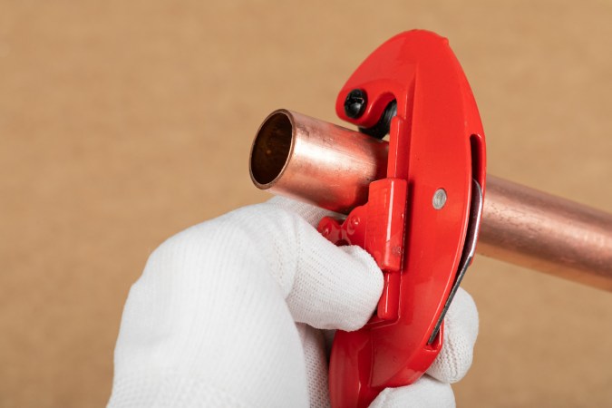 A person wearing a white glove is using a red pipe cutter to cut a copper pipe.