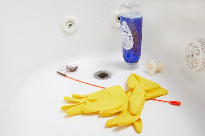 Rubber gloves, drain unclogger, and dish soap inside a white bathtub.