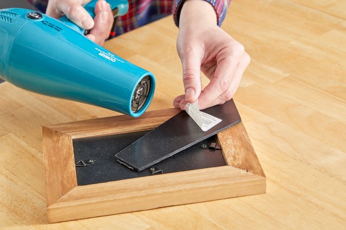 Woman uses a hair dryer to remove a sticker from a wood picture frame.