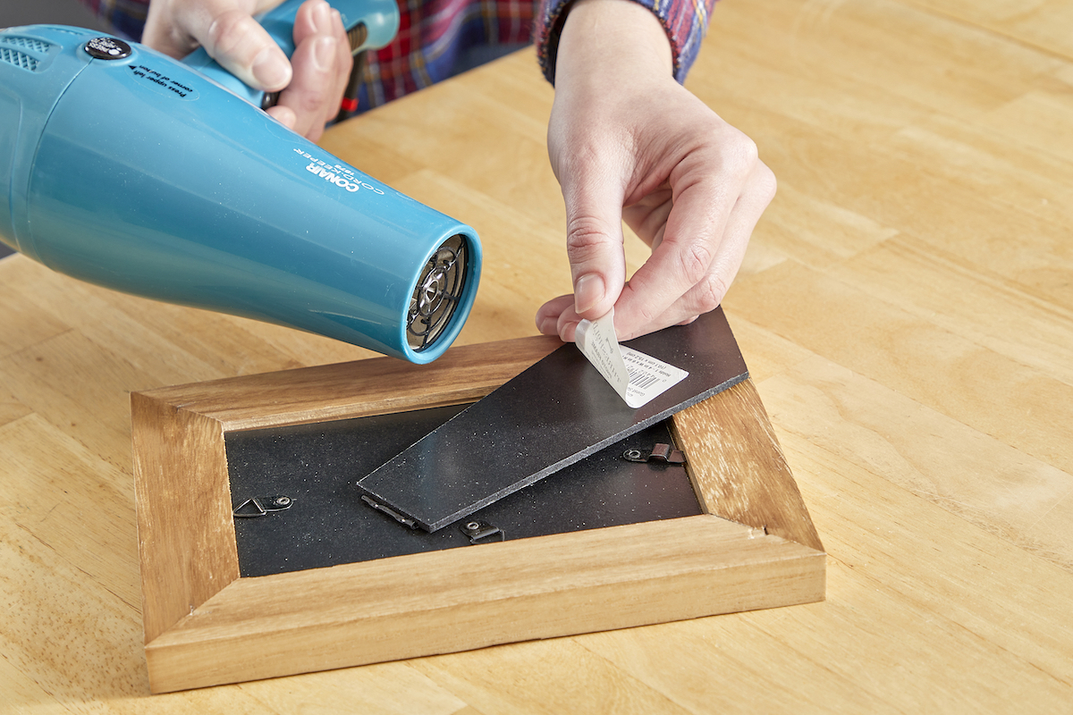 Woman uses a hair dryer to remove a sticker from a wood picture frame.