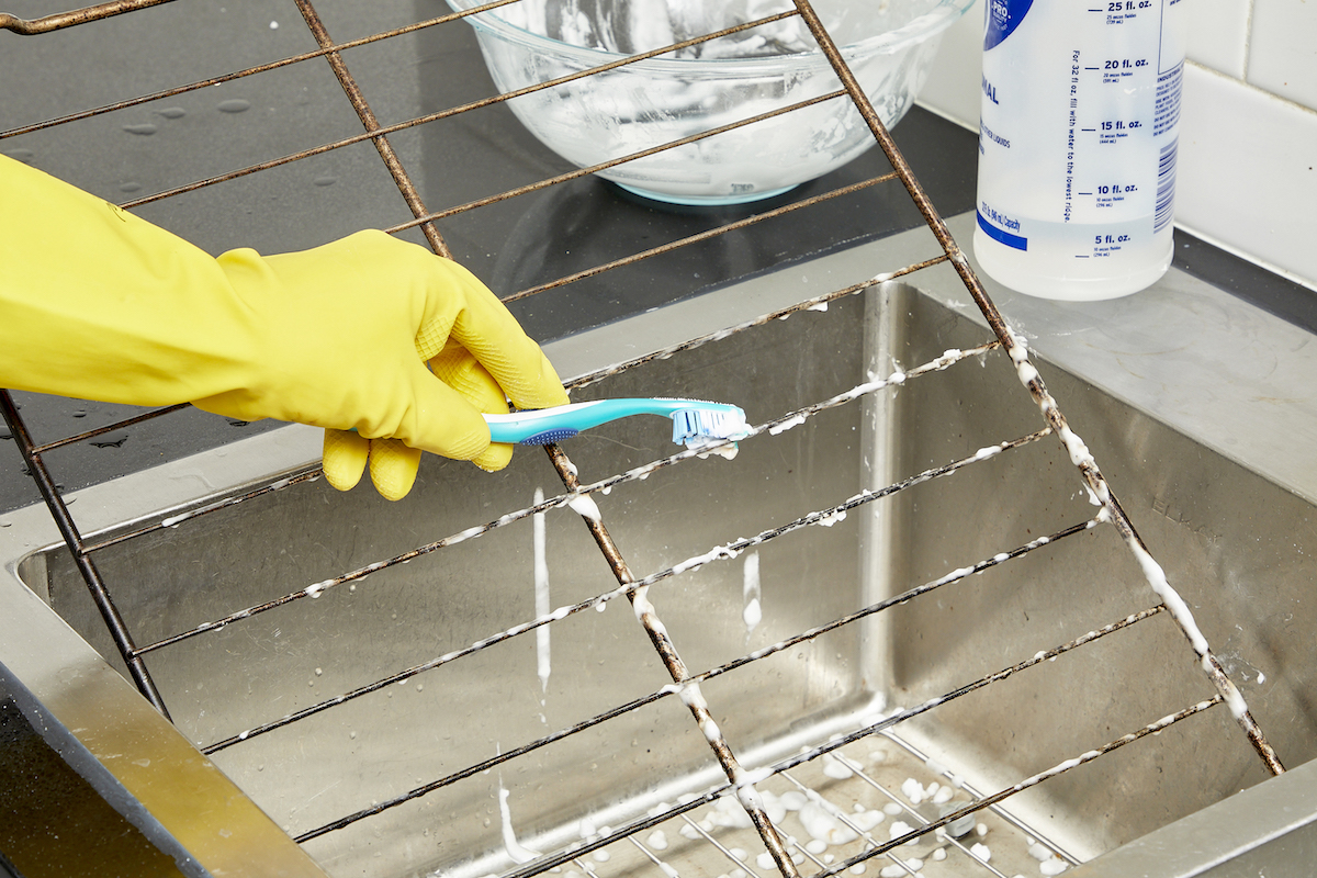 Woman wearing rubber gloves uses a toothbrush to scrub oven racks in a kitchen sink.