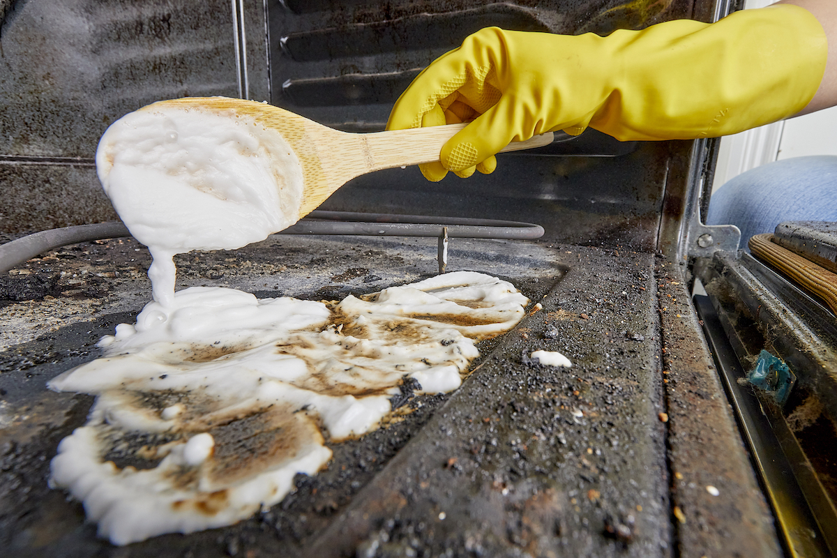 Woman uses a wooden spoon to spread homemade oven scrub inside a dirty oven.