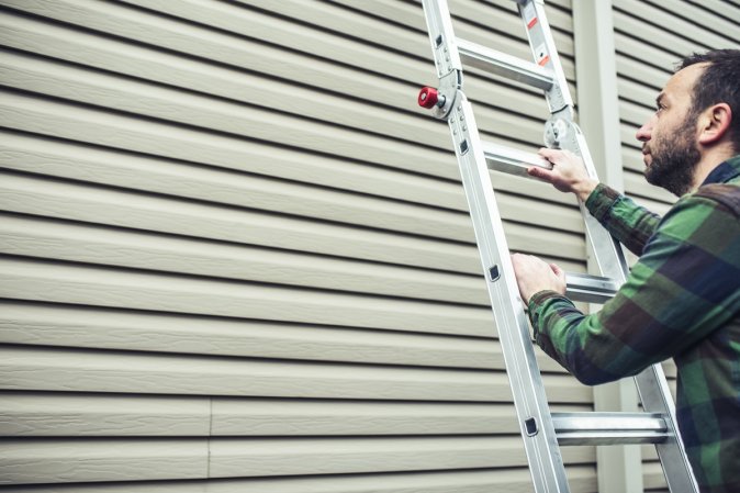 Repairman with green plaid shirt inspects tan home siding on a ladder.