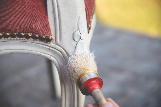 Person chalk painting wooden chair leg with white paint, pink chair cushion and brass nail heads visible.