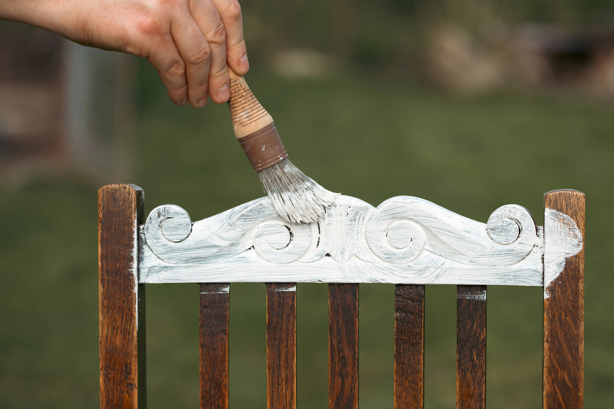 Man chalk painting the back of a scrolled wooden chair with white chalk paint. 