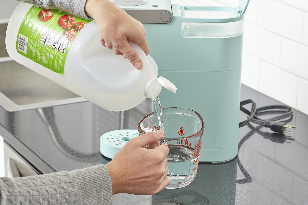 Woman pours vinegar from a gallon bottle into a measuring cup, in order to clean Keurig coffee maker in background.