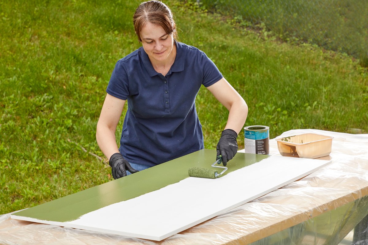 Woman in navy polo shirt uses a paint roller to paint a sheet of MDF green.