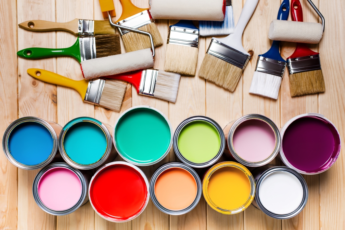 Multiple buckets of colored paints lined up next to a pile of paint brushes and rollers on a wooden floor