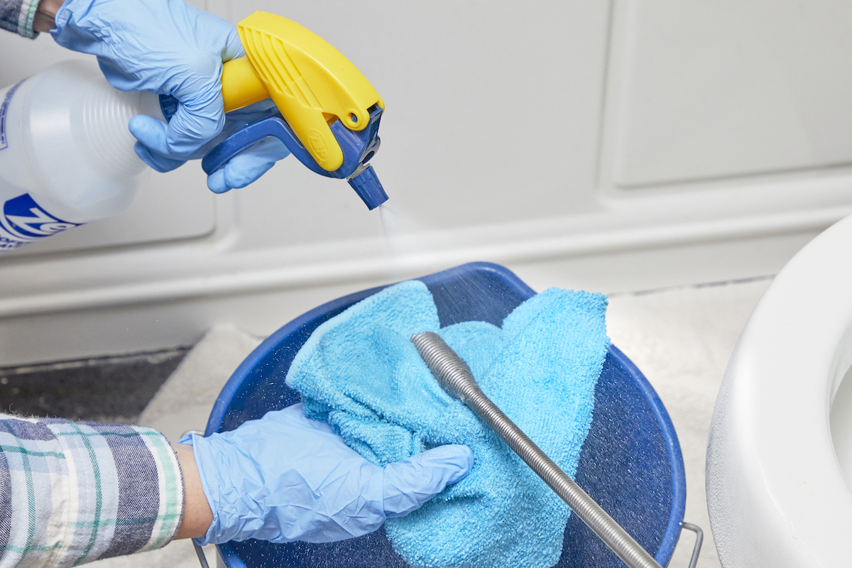 Person cleans a toilet auger by spraying it with a spray bottle and microfiber cloth held over a bucket.