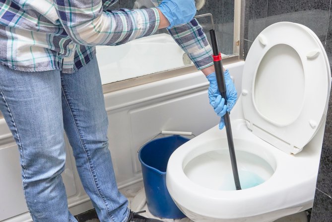 Person wearing gloves leans over toilet bowl with drain snake to clear clog.