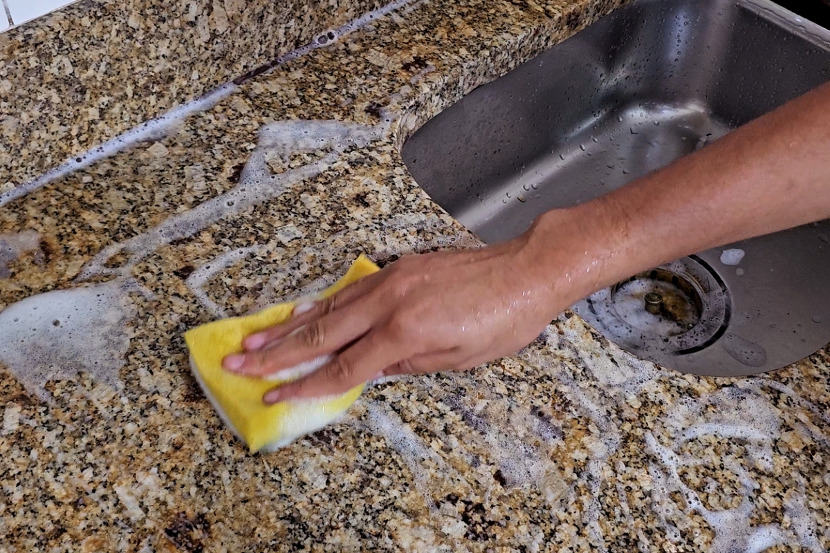 A person using a sponge and soap to clean granite countertop near sink.