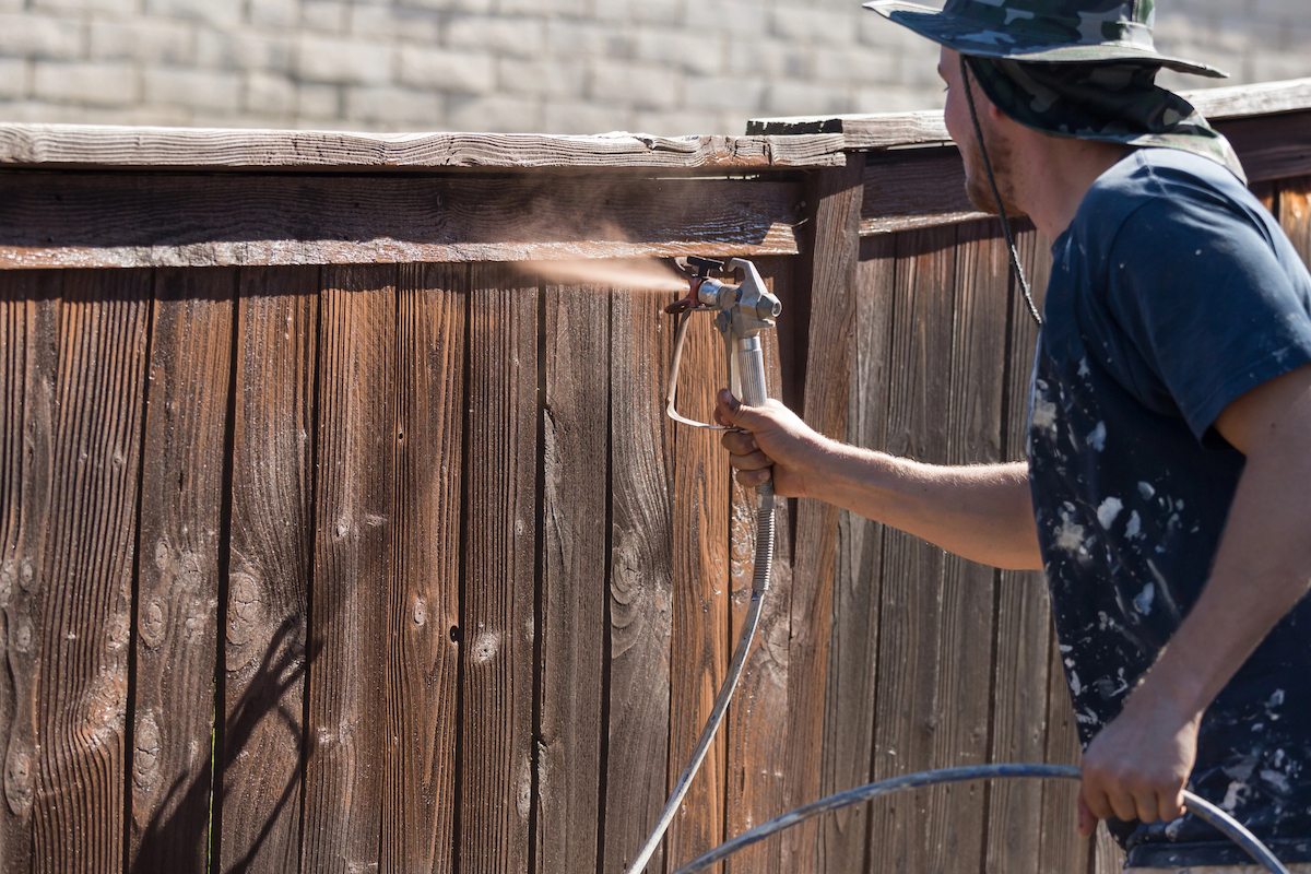 A person is using a paint sprayer to apply a second coat of wood stain to a wooden fence.