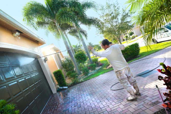 Man wearing a baseball cap pressure washes a brown garage door, palm trees in the background.