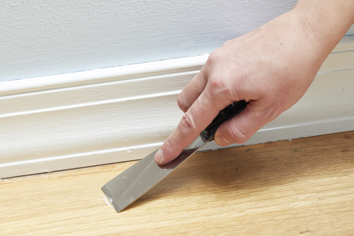 Woman uses a putty knife to scrape a bit of finish from a hardwood floor.