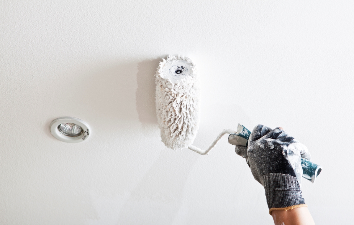 Gloved hand using roller to paint ceiling.