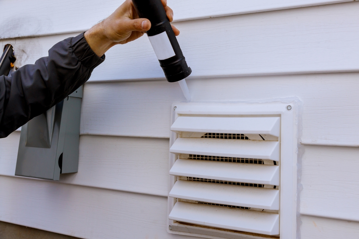 A person's arm is seen using caulk to seal the exterior vent to a home.