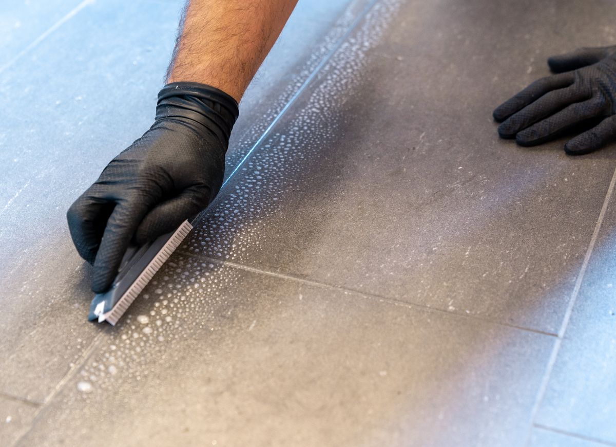 Cleaning grout lines on grey tiles using a scrub brush and gloves.