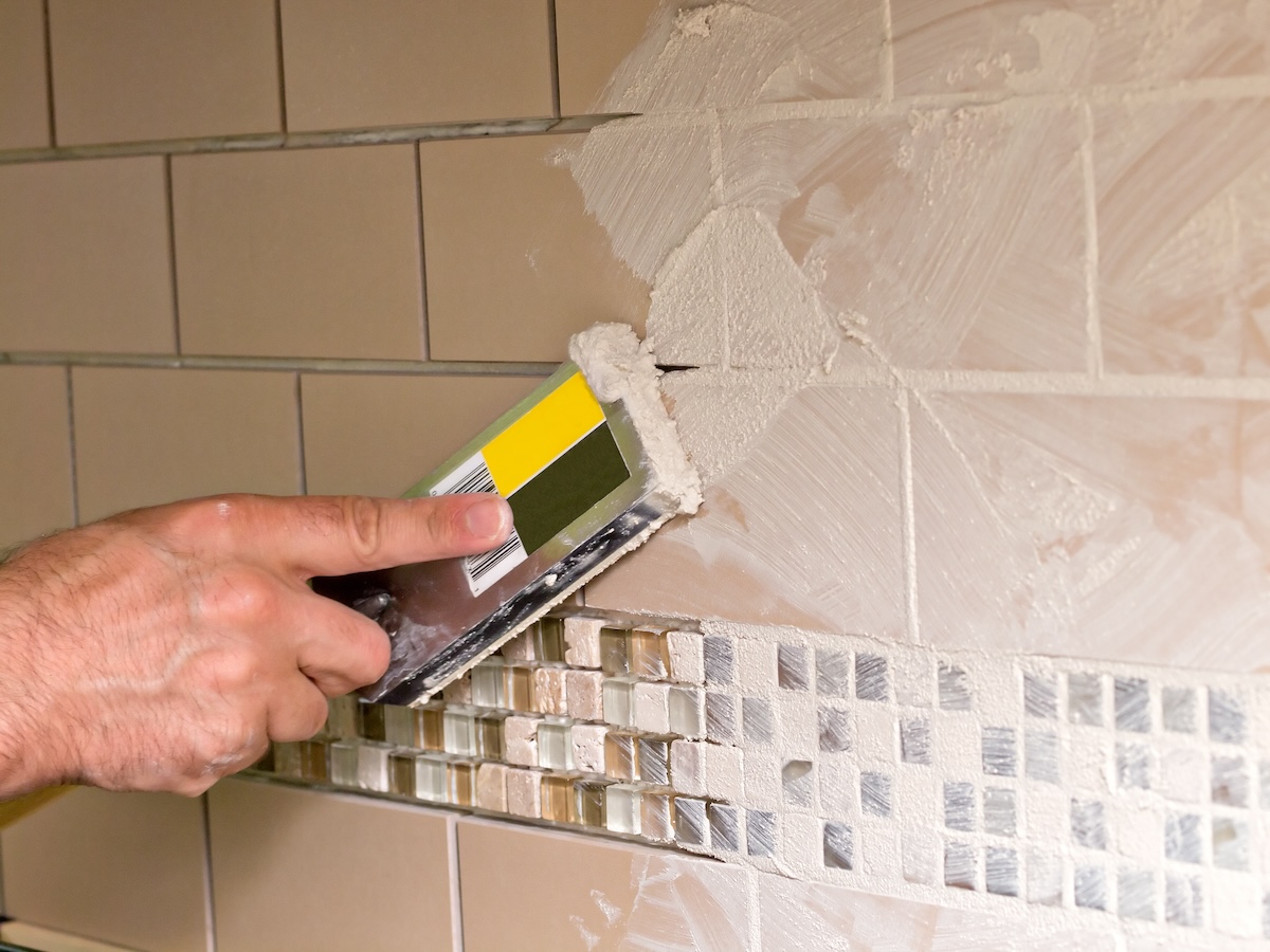 Hand using scrape to spread grout on beige tiles.