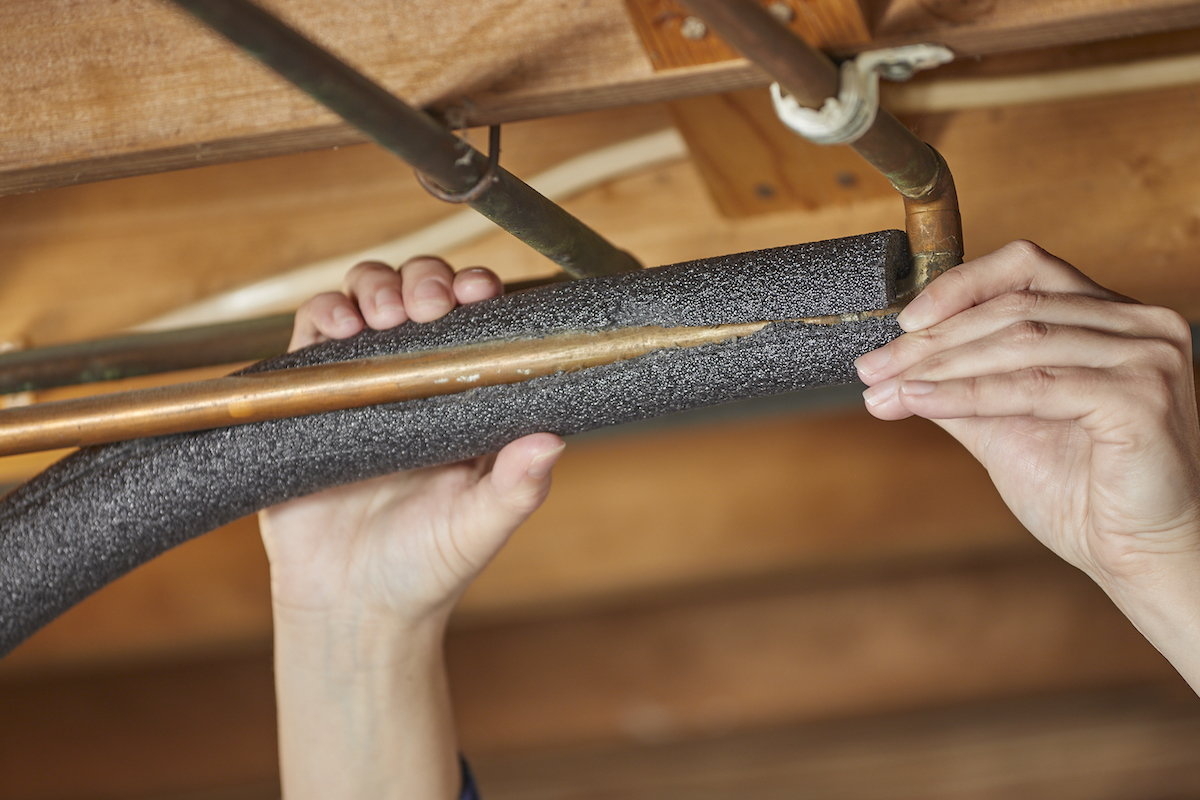 Woman insulates copper pipe along rafters in basement.