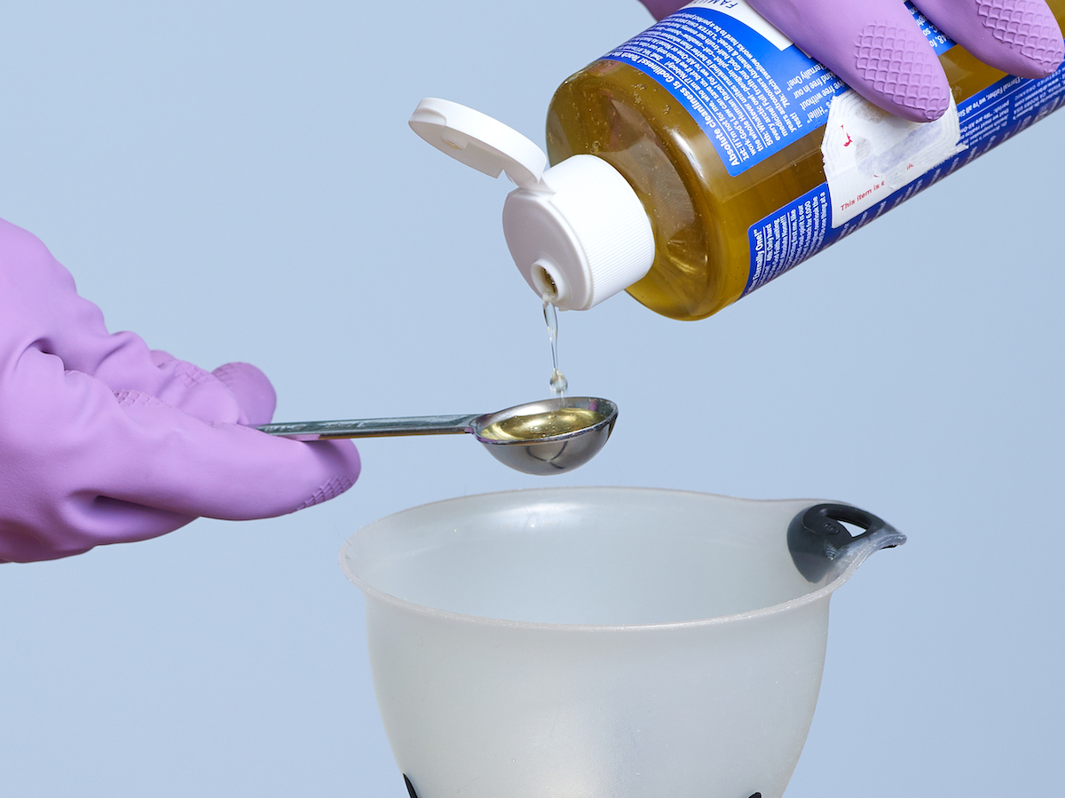 Woman wearing purple gloves measures castile soap into a funnel.