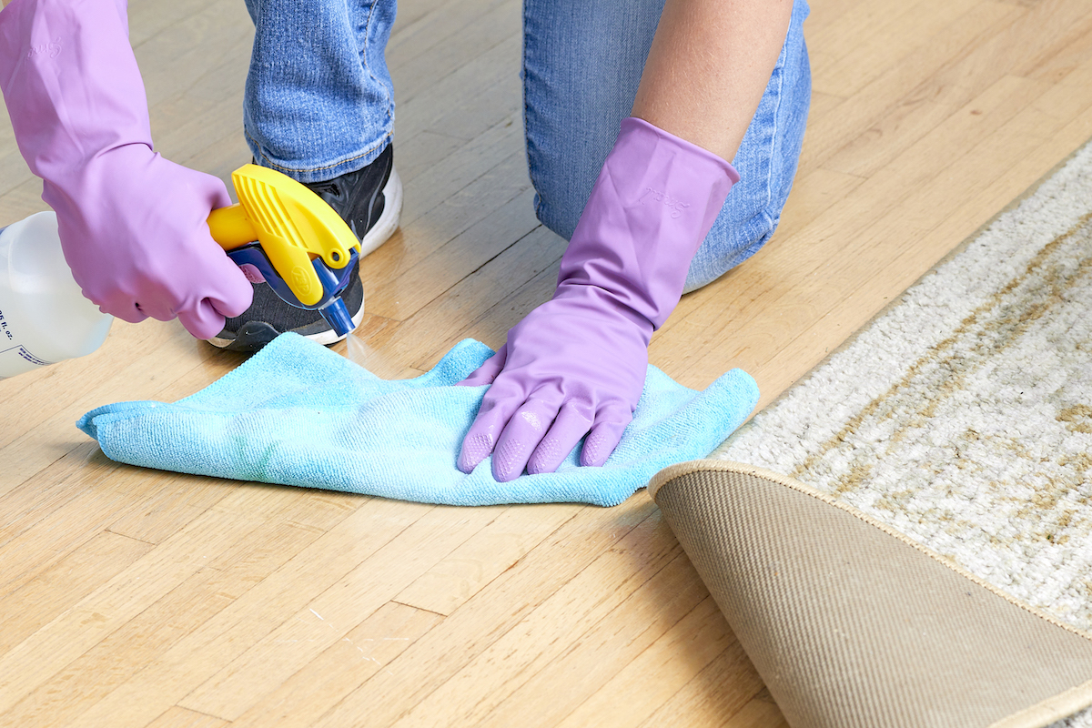 Woman sprays hardwood floor under an area rug, and wipes with a microfiber cloth.